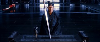 Movie still from “Hero” (2002), directed by Yimou Zhang – A man in a black robe holding a sword and standing in front of a statue; Medium shot, Low angle