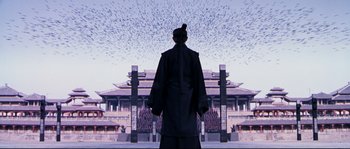 Movie still from “Hero” (2002), directed by Yimou Zhang – A man standing in front of a building with birds flying overhead; Extreme Wide shot, Low angle