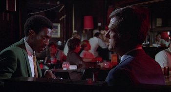Movie still from “Hickey & Boggs” (1972), directed by Robert Culp – Two men sitting at a table in front of a group of people; Medium shot, Over the shoulder angle