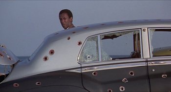 Movie still from “Hickey & Boggs” (1972), directed by Robert Culp – A man standing inside of a car with bullet holes in it; Medium shot, Low angle
