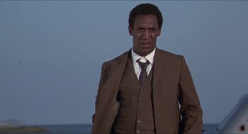 Movie still from “Hickey & Boggs” (1972), directed by Robert Culp – A man in a suit and tie standing in front of a body of water; Medium shot, Low angle
