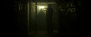 Movie still from “Hidden” (2015), directed by Ross Duffer – A man standing in a dark room with a light shining on him; Wide shot, Low angle