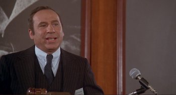 Movie still from “High Anxiety” (1977), directed by Mel Brooks – A man in a suit and tie standing in front of a microphone; Close Up shot, High angle