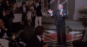 Movie still from “High Anxiety” (1977), directed by Mel Brooks – A man in a black suit is speaking into a microphone; Wide shot, High angle