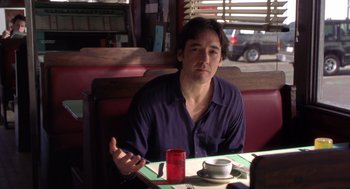 Movie still from “High Fidelity” (2000), directed by Stephen Frears – A man sitting at a table in front of a cup; Medium shot, Over the shoulder angle