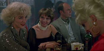 Movie still from “High Heels” (1991), directed by Pedro Almodóvar – A woman in a black dress sitting at a table with other people; Close Up shot, Over the shoulder angle