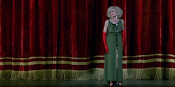 Movie still from “High Heels” (1991), directed by Pedro Almodóvar – An older woman wearing red gloves and a green dress; Wide shot, High angle