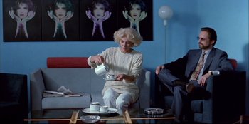 Movie still from “High Heels” (1991), directed by Pedro Almodóvar – An older woman sitting on a couch pouring tea into a cup; Wide shot, Over the shoulder angle