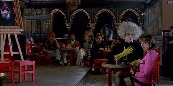 Movie still from “High Heels” (1991), directed by Pedro Almodóvar – A group of people sitting at tables in a room; Wide shot, Over the shoulder angle