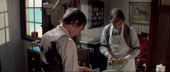 Movie still from “High Plains Drifter” (1973), directed by Clint Eastwood – A man in a barber shop cutting a man's hair; Medium shot, High angle