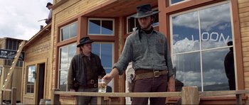 Movie still from “High Plains Drifter” (1973), directed by Clint Eastwood – A man holding a glass of beer while standing next to another man; Medium shot, Low angle