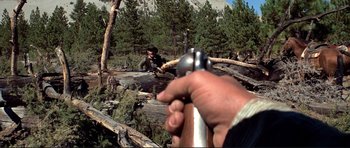 Movie still from “High Plains Drifter” (1973), directed by Clint Eastwood – A man is holding a gun in his hand while another man is sitting on a log; Wide shot, High angle