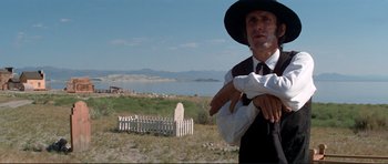 Movie still from “High Plains Drifter” (1973), directed by Clint Eastwood – A man wearing a black hat standing next to a white picket fence; Medium shot, Low angle