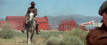 Movie still from “High Plains Drifter” (1973), directed by Clint Eastwood – A man riding a horse in front of a red building; Wide shot, Low angle
