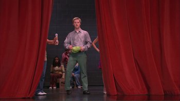 Movie still from “High School Musical” (2006), directed by Kenny Ortega – A man standing on a stage holding a bowl of fruit; Wide shot, Low angle