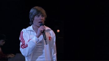 Movie still from “High School Musical” (2006), directed by Kenny Ortega – A man in white jacket holding a microphone; Medium shot, Over the shoulder angle
