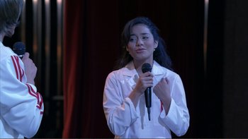 Movie still from “High School Musical” (2006), directed by Kenny Ortega – A woman holding a microphone while standing on a stage; Medium shot, Over the shoulder angle