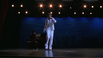Movie still from “High School Musical” (2006), directed by Kenny Ortega – A man standing on a stage holding a microphone; Wide shot, Low angle