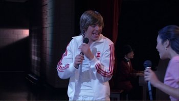 Movie still from “High School Musical” (2006), directed by Kenny Ortega – A person holding a microphone on a stage; Medium shot, Over the shoulder angle