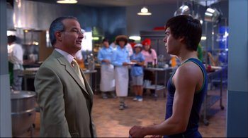 Movie still from “High School Musical 2” (2007), directed by Kenny Ortega – A man and a woman standing in front of a group of people in a kitchen; Medium shot, Over the shoulder angle