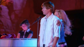 Movie still from “High School Musical 2” (2007), directed by Kenny Ortega – A young man standing in front of a microphone with two other young women; Medium shot, Over the shoulder angle