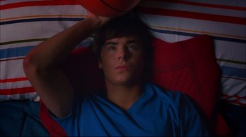 Movie still from “High School Musical 2” (2007), directed by Kenny Ortega – A man laying in a bed with a basketball on top of his head; Medium shot, Overhead angle