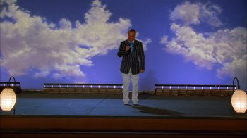Movie still from “High School Musical 2” (2007), directed by Kenny Ortega – A man standing on a stage in front of a blue sky; Wide shot, Low angle