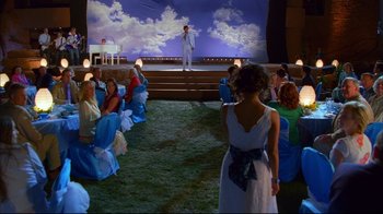 Movie still from “High School Musical 2” (2007), directed by Kenny Ortega – A group of people sitting in a field watching a man on a stage; Extreme Wide shot, Over the shoulder angle
