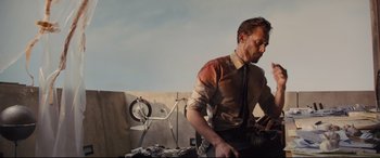 Movie still from “High-Rise” (2015), directed by Ben Wheatley – A man standing in front of a table with papers on top of it; Medium shot, Low angle