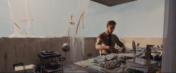 Movie still from “High-Rise” (2015), directed by Ben Wheatley – A man sitting at a table with papers on it; Medium shot, Low angle