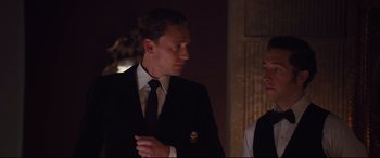 Movie still from “High-Rise” (2015), directed by Ben Wheatley – A man in a suit and tie standing next to a woman in a black suit; Medium shot, Over the shoulder angle