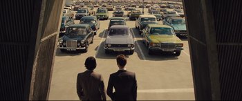 Movie still from “High-Rise” (2015), directed by Ben Wheatley – Two men are standing in a parking lot looking at many parked cars; Wide shot, High angle