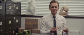 Movie still from “High-Rise” (2015), directed by Ben Wheatley – A man sitting at a desk holding a pencil; Medium shot, Over the shoulder angle