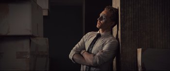 Movie still from “High-Rise” (2015), directed by Ben Wheatley – A man with his arms crossed leaning up against a wall; Medium shot, Low angle
