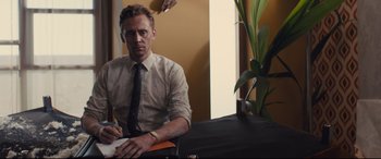 Movie still from “High-Rise” (2015), directed by Ben Wheatley – A man sitting at a table writing on a piece of paper; Medium shot, Low angle
