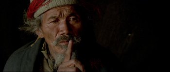 Movie still from “Himalaya” (1999), directed by Eric Valli – An old man with a beard and a hat is holding his finger to his lips; Close Up shot, High angle
