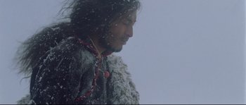 Movie still from “Himalaya” (1999), directed by Eric Valli – A man with long black hair and a beard standing in the snow; Close Up shot, Low angle