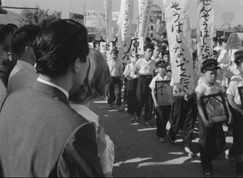 Movie still from “Hiroshima Mon Amour” (1959), directed by Alain Resnais – A group of people standing next to each other on a street; Medium shot, Over the shoulder angle