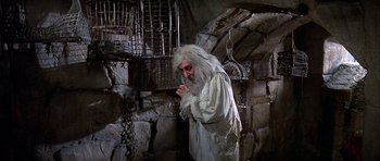 Movie still from “History of the World: Part I” (1981), directed by Mel Brooks – An old man with long white hair and a white beard standing next to a stone wall; Medium shot, High angle