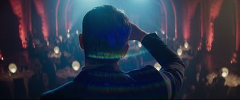 Movie still from “Hitman's Wife's Bodyguard” (2021), directed by Patrick Hughes – A man is sitting in front of a crowd at an event; Medium shot, Over the shoulder angle