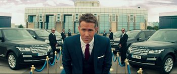 Movie still from “Hitman's Wife's Bodyguard” (2021), directed by Patrick Hughes – A man wearing a suit and tie standing in front of a group of cars; Wide shot, Over the shoulder angle