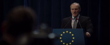 Movie still from “Hitman's Wife's Bodyguard” (2021), directed by Patrick Hughes – A man standing at a podium with a microphone; Medium shot, Over the shoulder angle