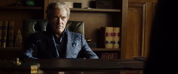 Movie still from “Hitman's Wife's Bodyguard” (2021), directed by Patrick Hughes – An older man sitting at a desk in a room; Medium shot, Over the shoulder angle