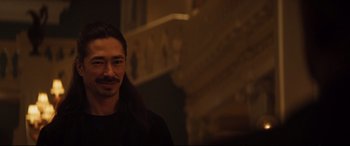 Movie still from “Hitman's Wife's Bodyguard” (2021), directed by Patrick Hughes – A man with long black hair standing in front of a staircase; Close Up shot, Over the shoulder angle