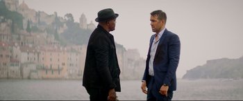 Movie still from “Hitman's Wife's Bodyguard” (2021), directed by Patrick Hughes – Two men standing next to each other near a body of water; Medium shot, Over the shoulder angle