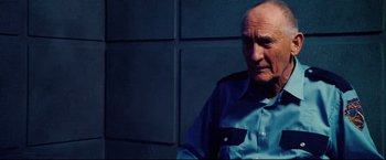 Movie still from “Hobo with a Shotgun” (2011), directed by Jason Eisener – An older man wearing a blue shirt is sitting in front of a blue wall; Close Up shot, Over the shoulder angle