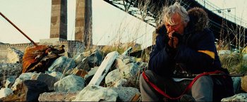 Movie still from “Hobo with a Shotgun” (2011), directed by Jason Eisener – An older man sitting on a rock near a pile of rocks; Medium shot, Low angle