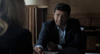 Movie still from “Holy Lands” (2017), directed by Amanda Sthers – A man in a suit sitting in front of a lamp; Medium shot, Over the shoulder angle