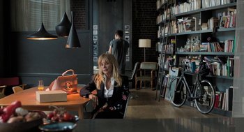 Movie still from “Holy Lands” (2017), directed by Amanda Sthers – A woman sitting at a table in front of bookshelves; Wide shot, High angle