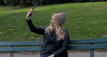 Movie still from “Holy Lands” (2017), directed by Amanda Sthers – A woman sitting on top of a bench taking a picture of herself; Medium shot, Over the shoulder angle